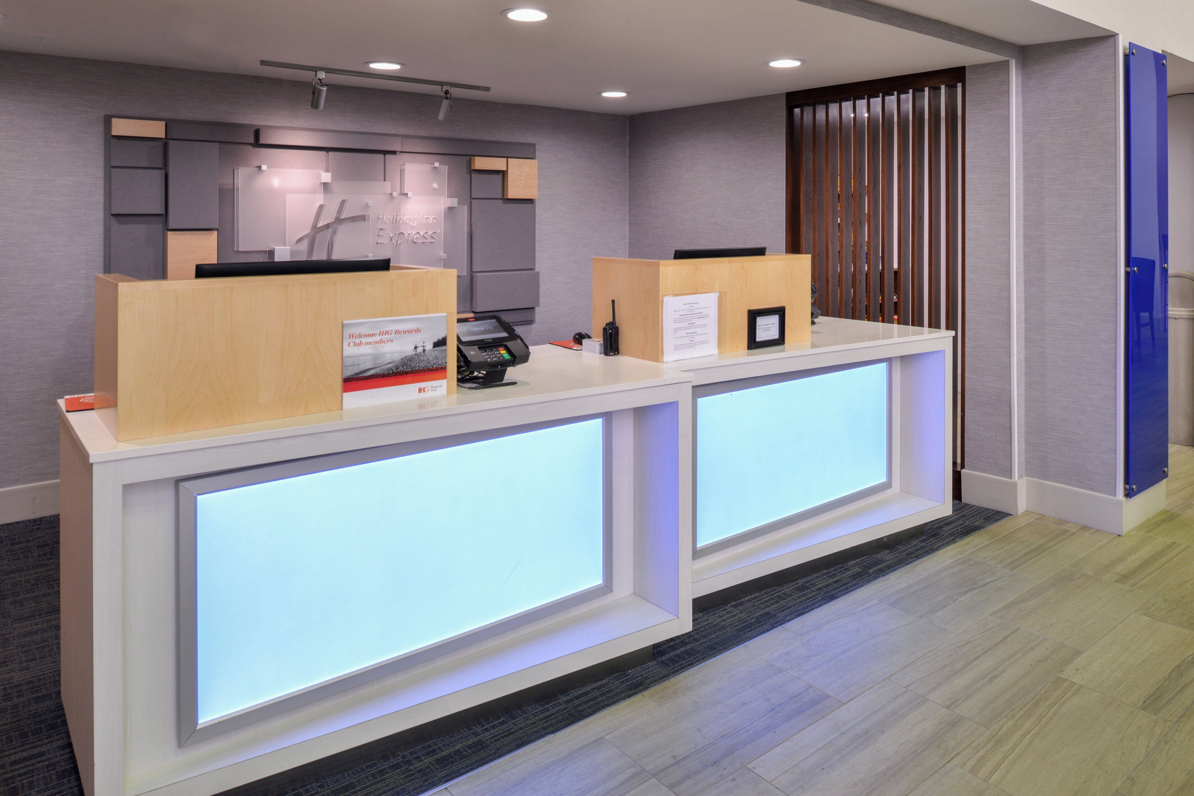 A reception desk with a modern design, featuring two counters with illuminated panels, a reception area with a wooden panel backdrop, and a blue door on the right.