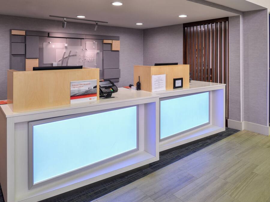 A reception desk with a modern design, featuring two counters with illuminated panels, a reception area with a wooden panel backdrop, and a blue door on the right.
