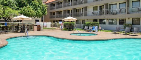 A hotel pool area with a clear pool, lounge chairs, umbrellas, and a multi-story building with balconies.
