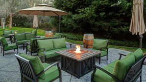 An outdoor patio area with green cushioned seating, a central fire pit, and wooden barrels.