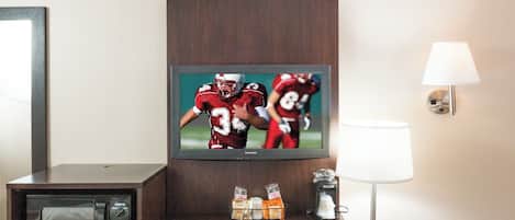 Une chambre d’hôtel équipée d’une télévision diffusant un événement sportif, d’un bureau avec une lampe, d’une chaise et d’un mini-bar.