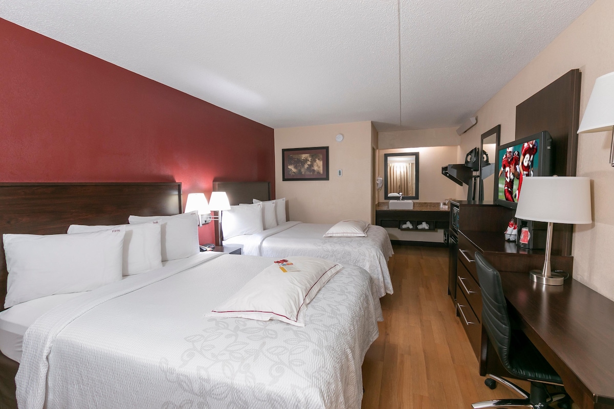 A hotel room with two beds, a desk, a chair, a lamp, a mirror, and a TV.