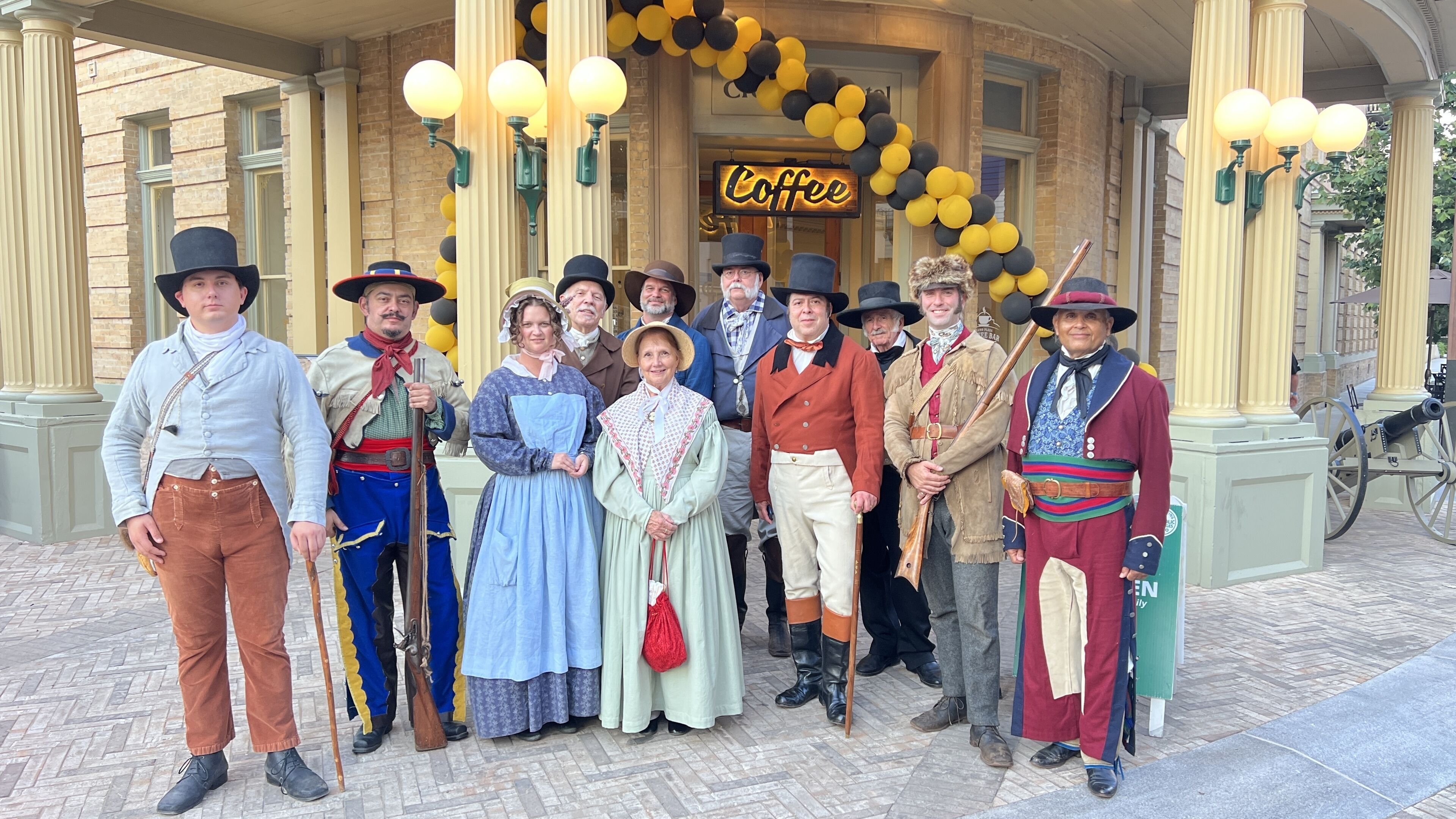 Un grupo de personas vestidas con trajes de época frente a un edificio con un letrero que dice 'Coffee' y globos negros y amarillos.