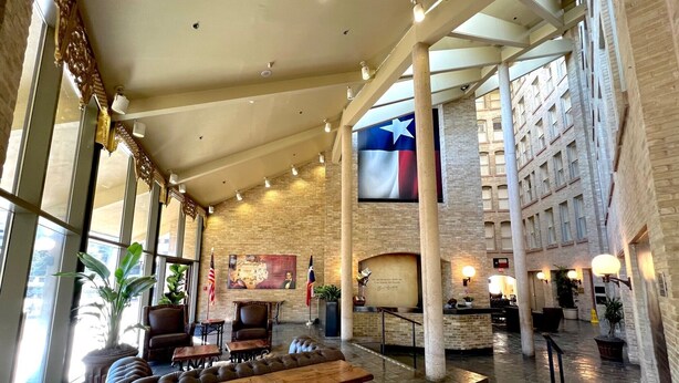 A spacious lobby with a high ceiling, large windows, and a Texas flag. There are seating areas with tables and chairs, and a large screen displaying a map. - Crockett Hotel (San Antonio)