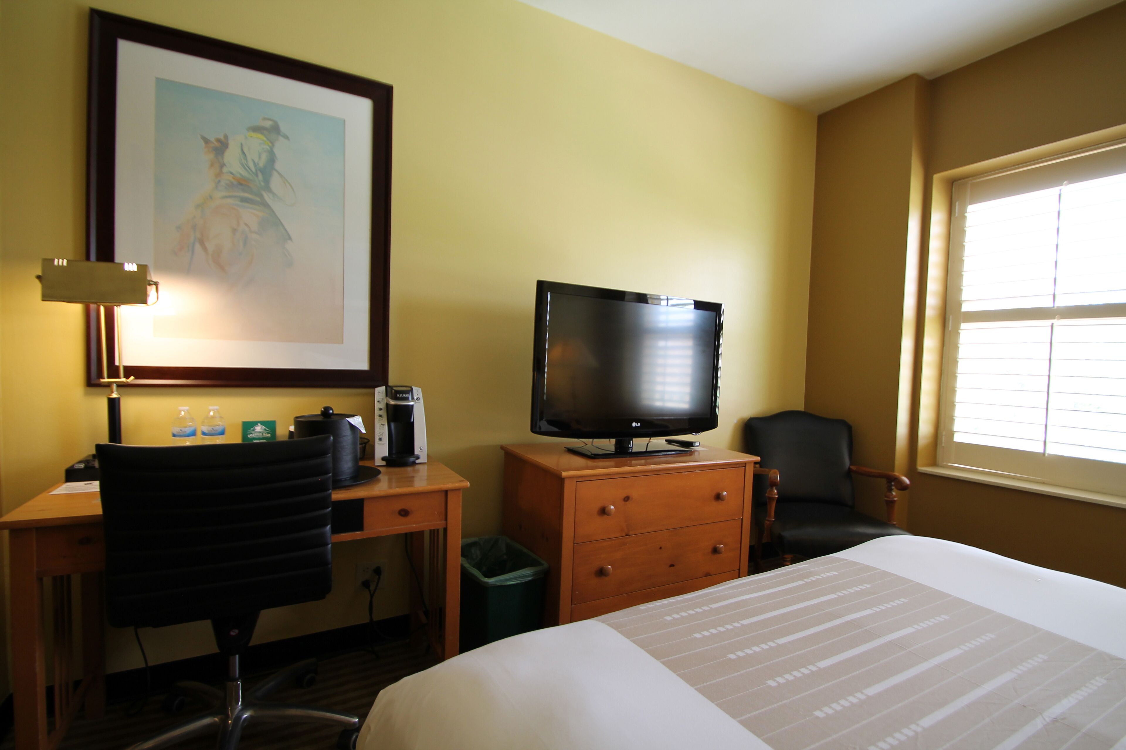 Habitación de hotel con cama, escritorio con silla, televisor, un cuadro en la pared y ventana con persianas.