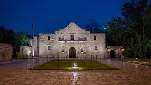 A historic stone building with a central arched entrance, a flagpole, and a well-maintained lawn in front. - Crockett Hotel (San Antonio)