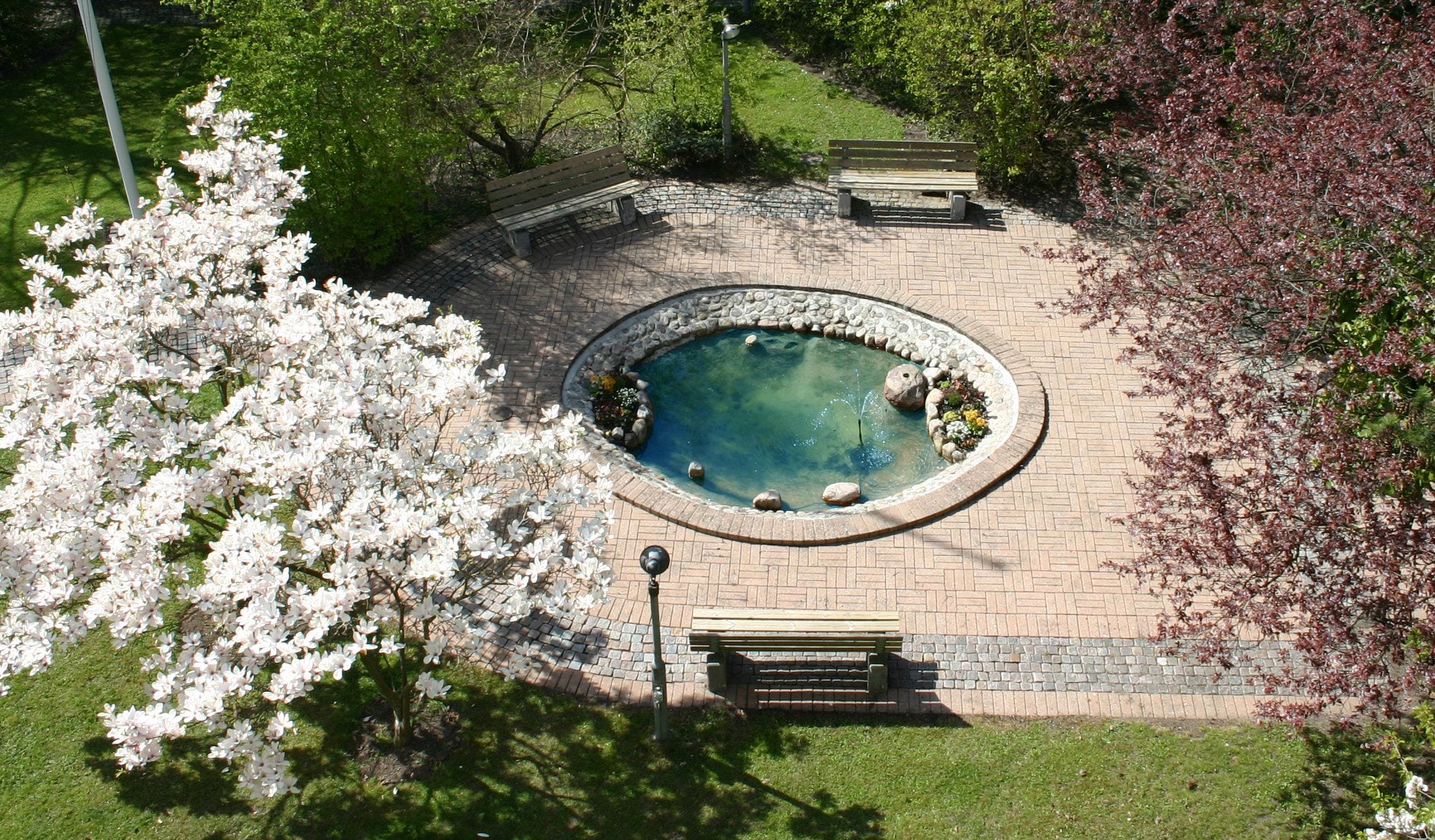 A circular stone pool with a fountain, surrounded by a brick pathway and benches.