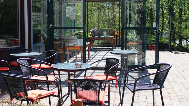 An outdoor patio with round tables and wicker chairs, surrounded by glass walls offering views of trees and a garden. - Hotel Djingis Khan (Lund)