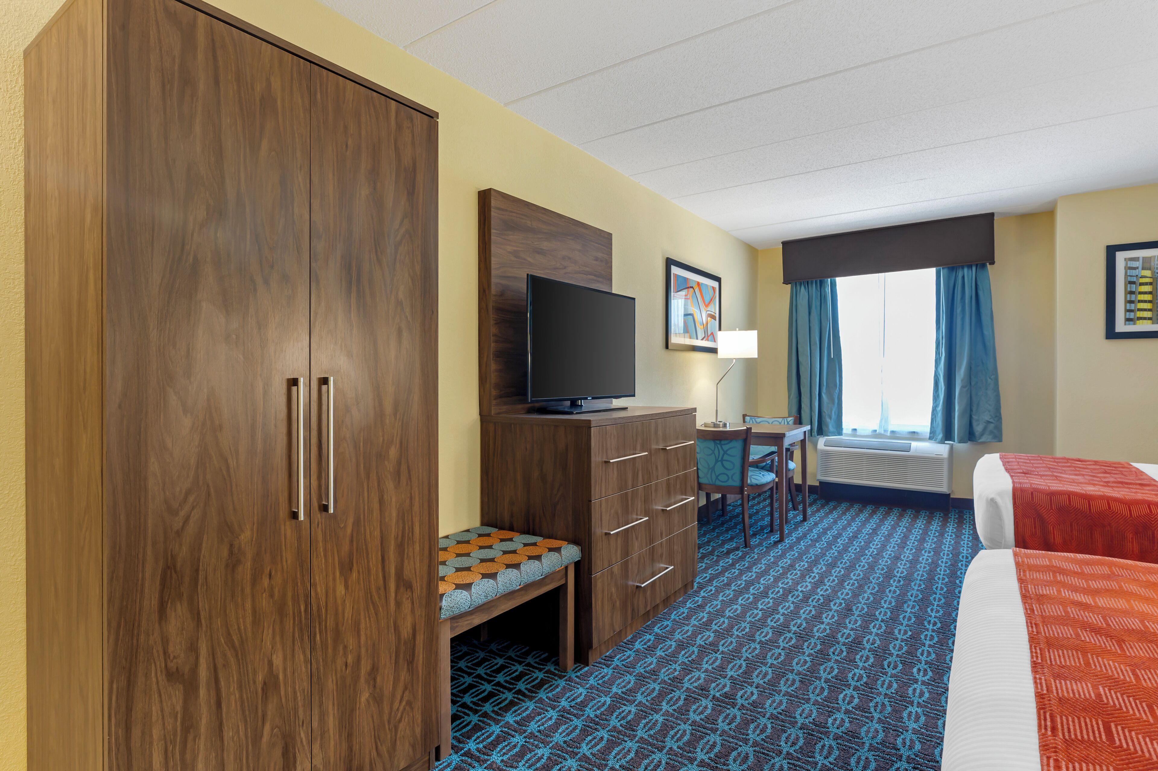 A hotel room with a large wardrobe, a television, a desk with a chair, a bed, and a window with curtains.