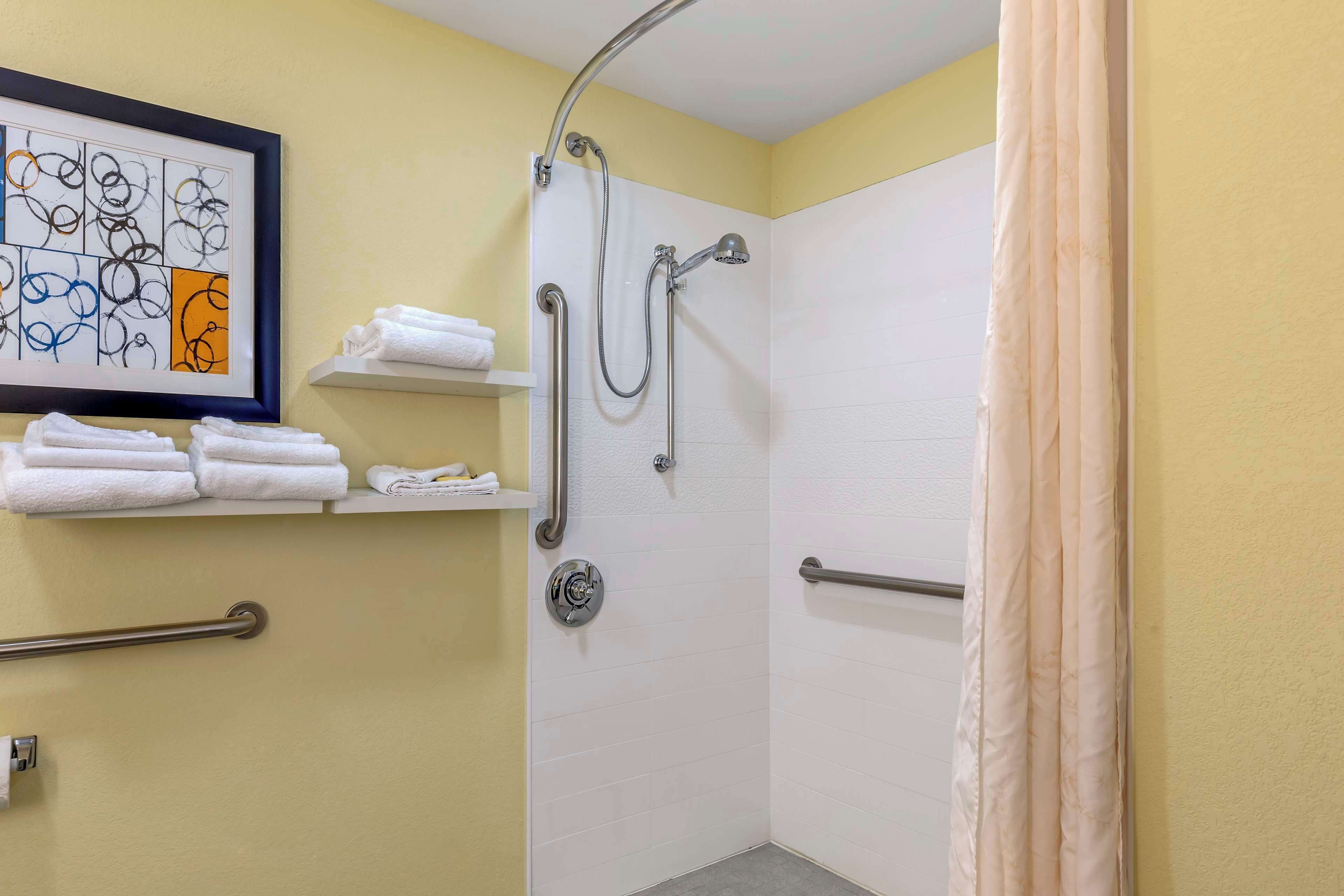 Un espace douche équipé d’une barre d’appui, d’un porte-serviettes avec des serviettes pliées, et d’une œuvre d’art abstraque encadrée accrochée au mur.