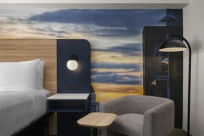 Ein Hotelzimmer mit Bett, einem kleinen Tisch, einem Stuhl, einer Wandlampe und einem Sonnenuntergangsmural.