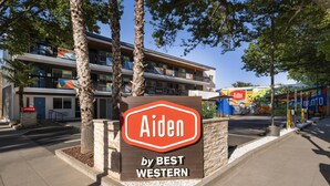 A hotel sign with 'Aiden by BEST WESTERN' and a modern building with balconies and a parking area.