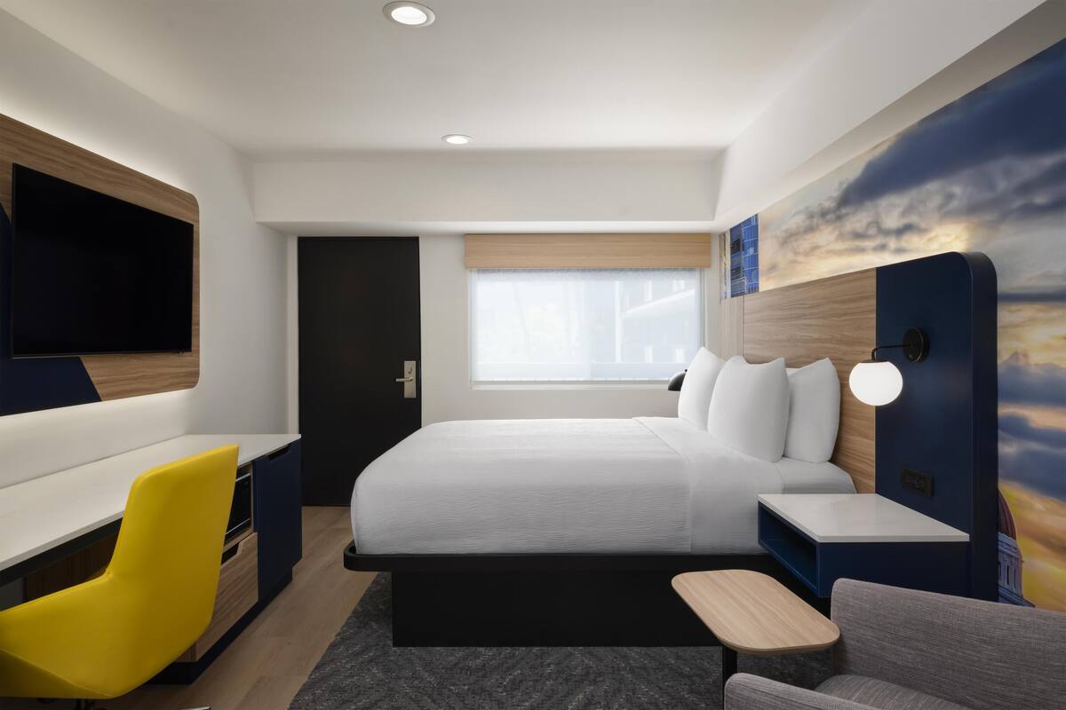 A modern hotel room with a large bed, a desk, a chair, and a TV.