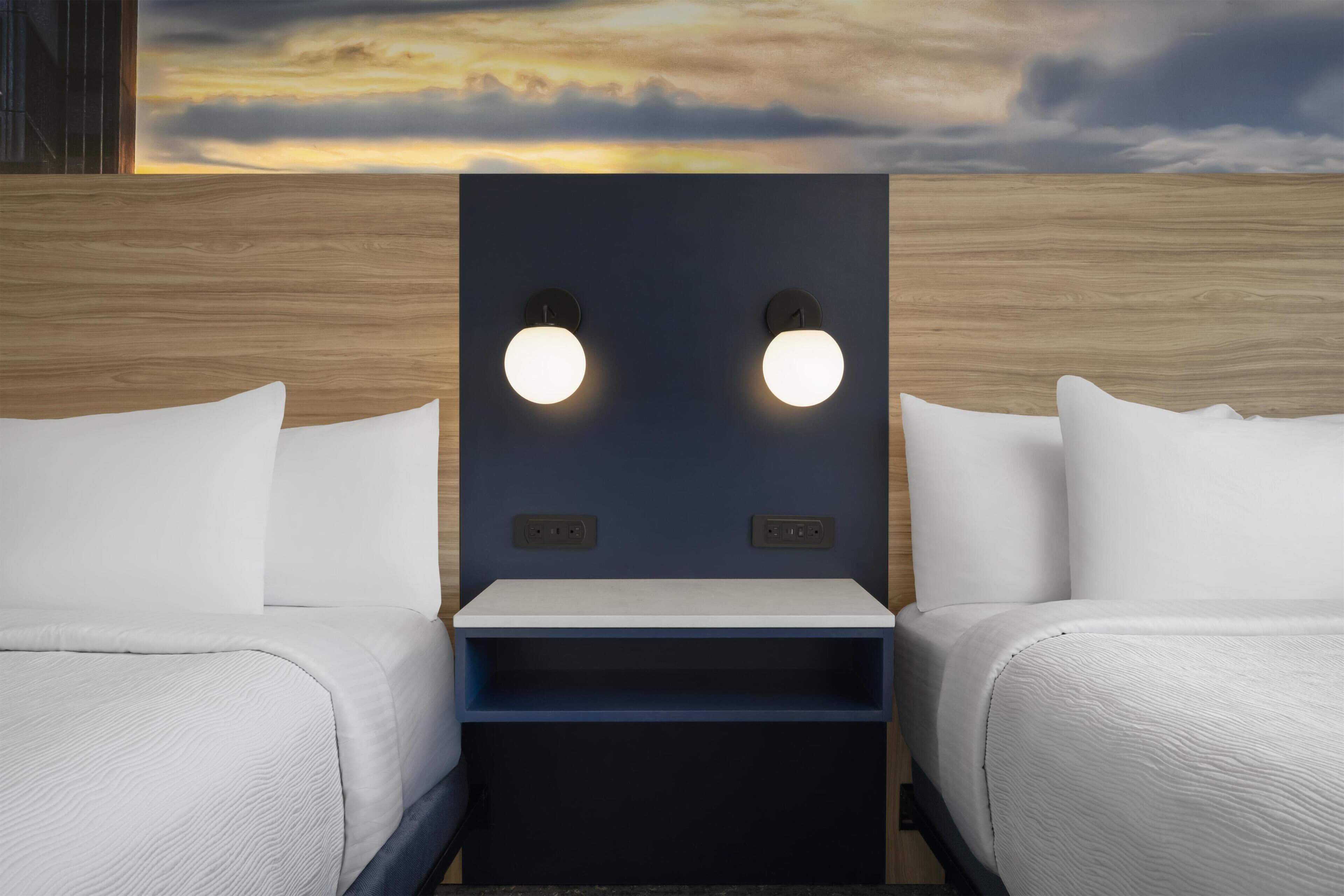 A hotel room with two beds, a nightstand, wall-mounted reading lights, and a view of the sky.