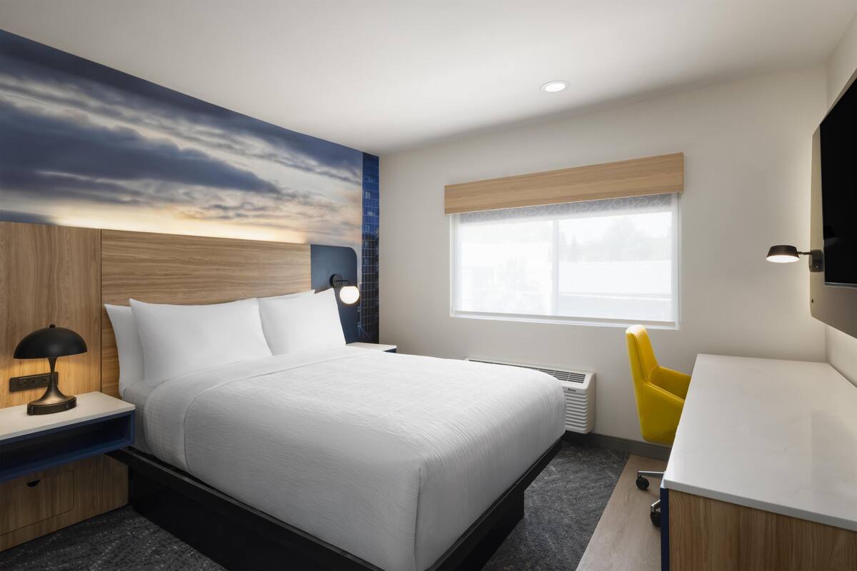 A modern hotel room with a bed, bedside table, lamp, and a window with blinds.