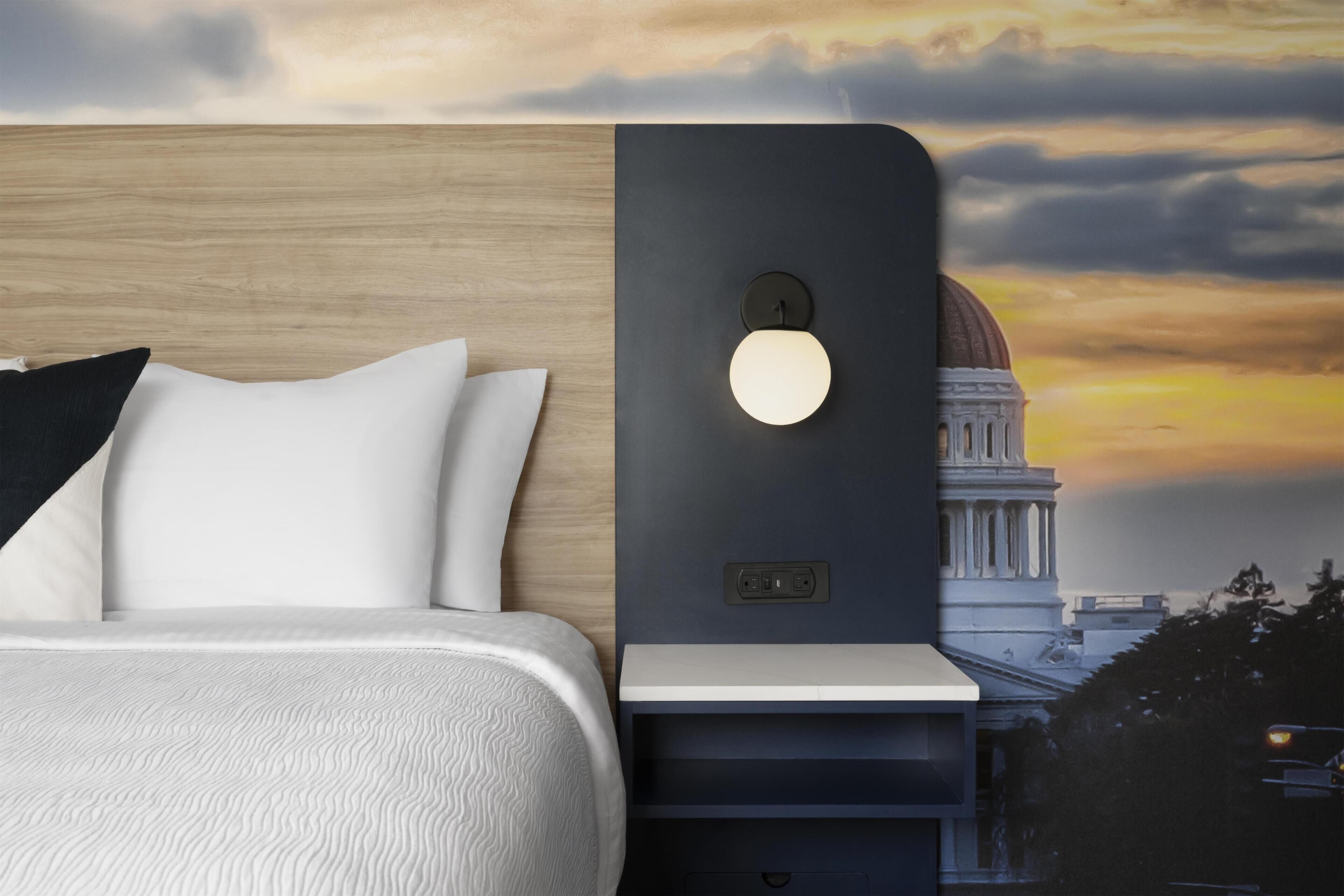 A bed with white bedding, a nightstand with a wall-mounted light, and a view of a building with a dome.