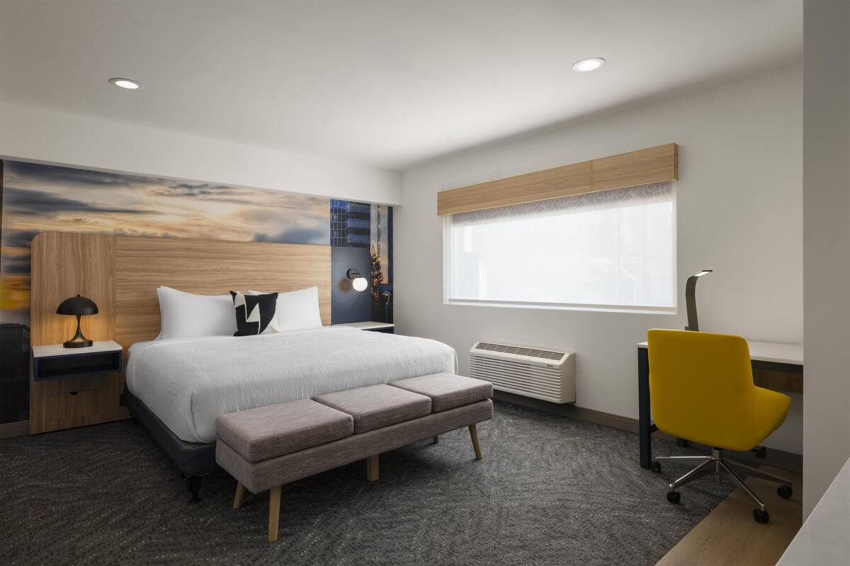 A modern hotel room with a large bed, a desk, a chair, and a large window.