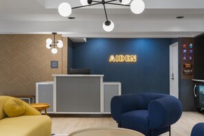 Ein Empfangsbereich mit blauer Wand, einer Rezeption, einem gelben Sofa, einem blauen Sessel und einer Deckenleuchte.
