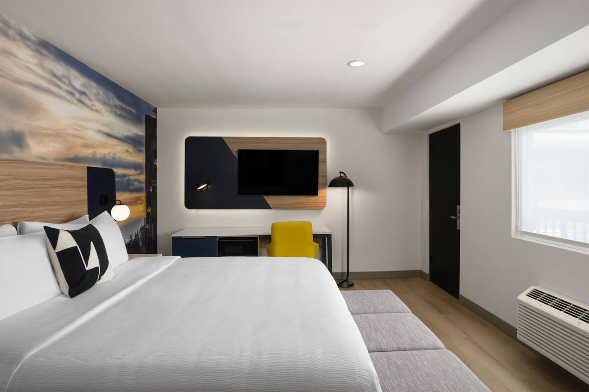 A modern hotel room with a large bed, a desk, a flat-screen TV, and a large window.