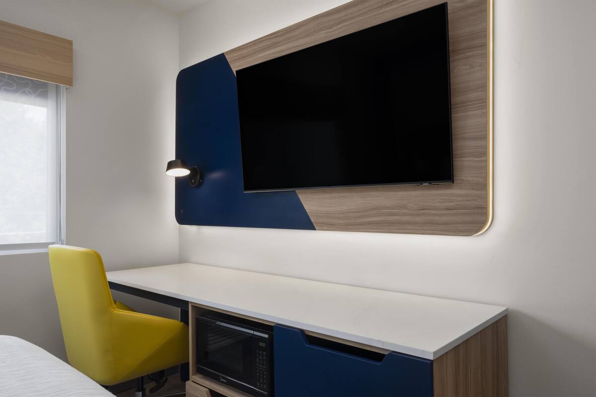 A modern room with a flat-screen TV mounted on a wall, a desk with a microwave, and a yellow chair.