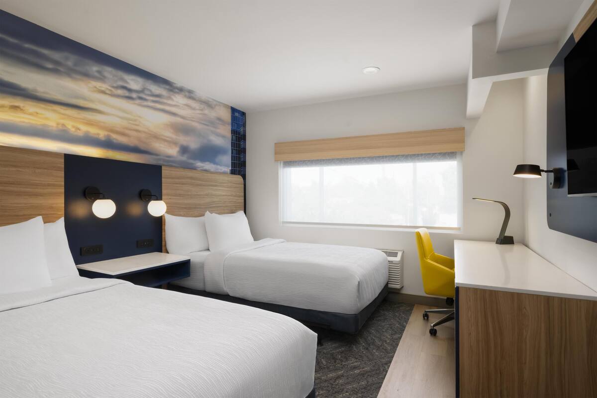 A hotel room with two beds, a desk, a chair, a TV, and a large window.