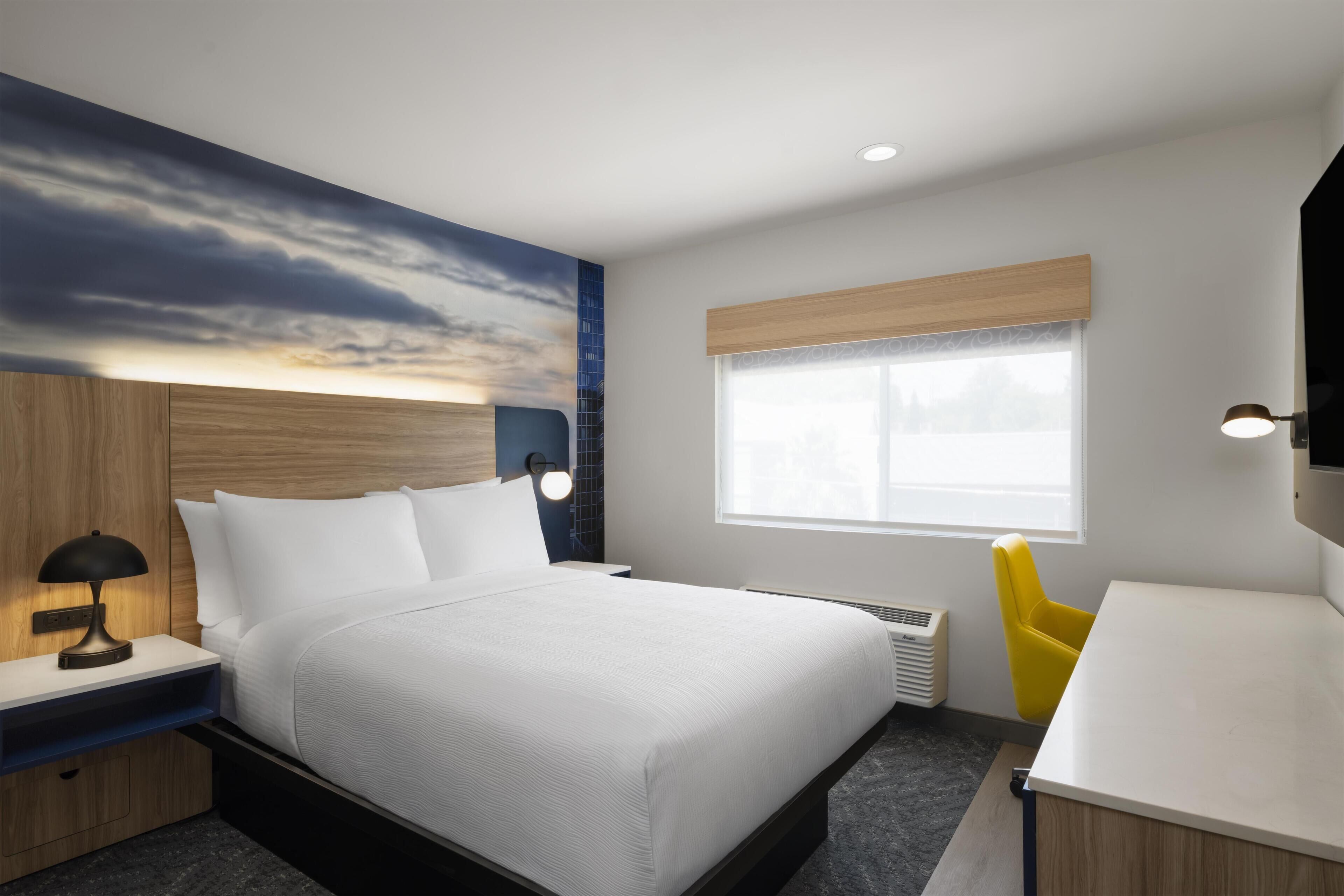 A hotel room with a bed, bedside table, lamp, window, and a yellow chair.