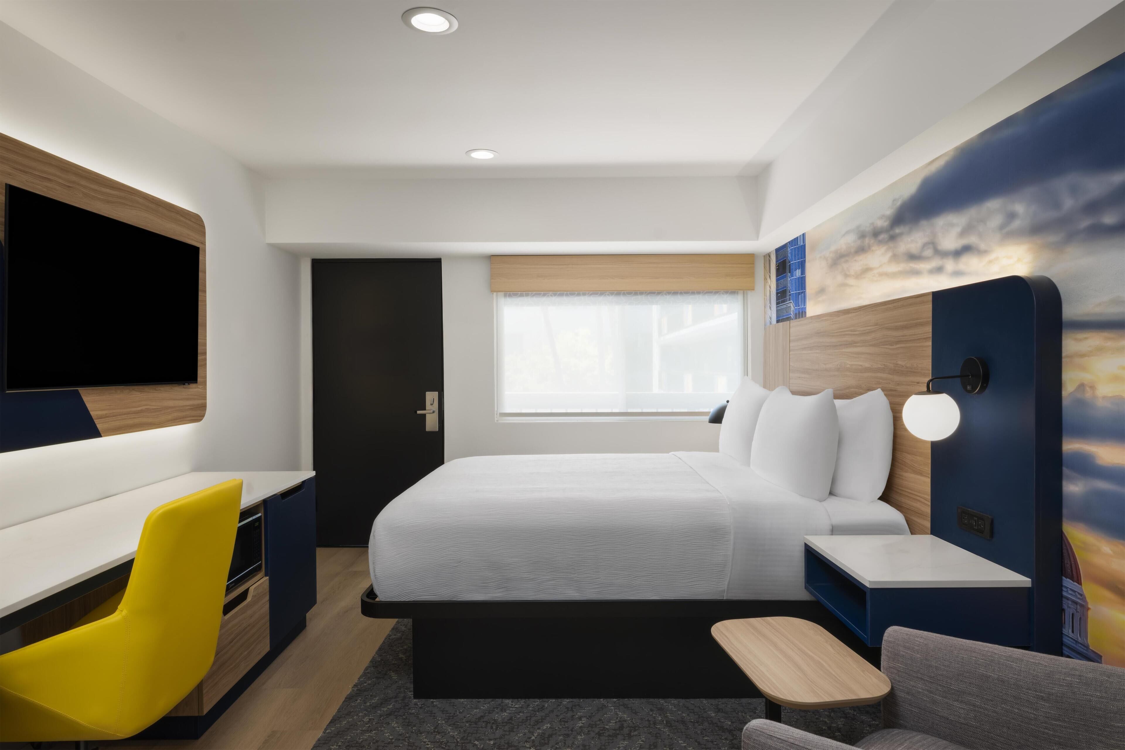 A modern hotel room with a bed, a desk, a chair, a TV, and a wall with a sunset mural.