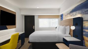 A modern hotel room with a bed, a desk, a chair, a TV, and a wall with a sunset mural.