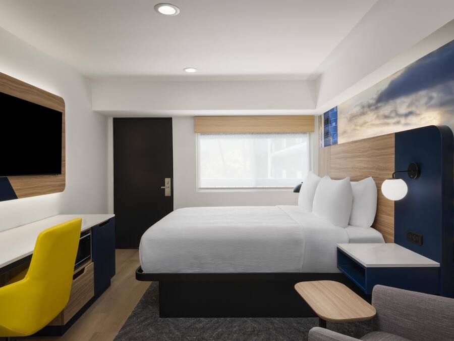 A modern hotel room with a bed, a desk, a chair, a TV, and a wall with a sunset mural.