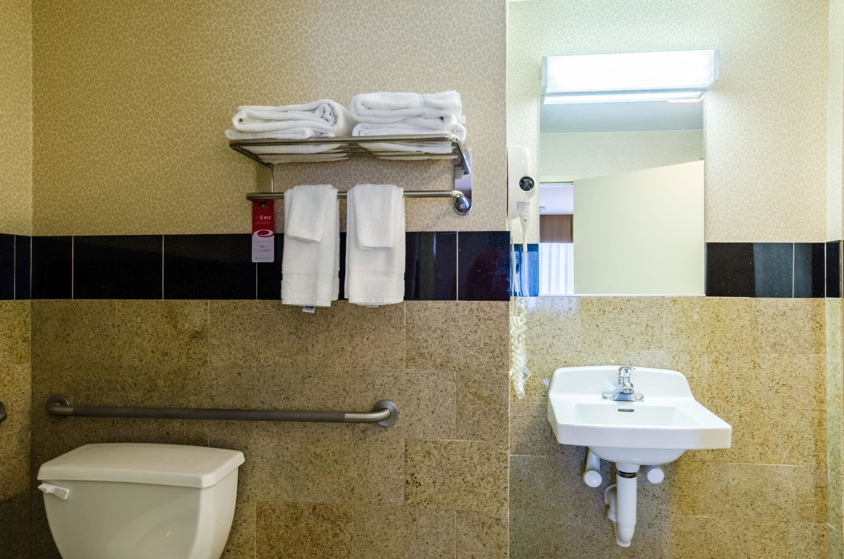 A bathroom with a toilet, a sink, a towel rack with folded towels, a handrail, and a mirror.