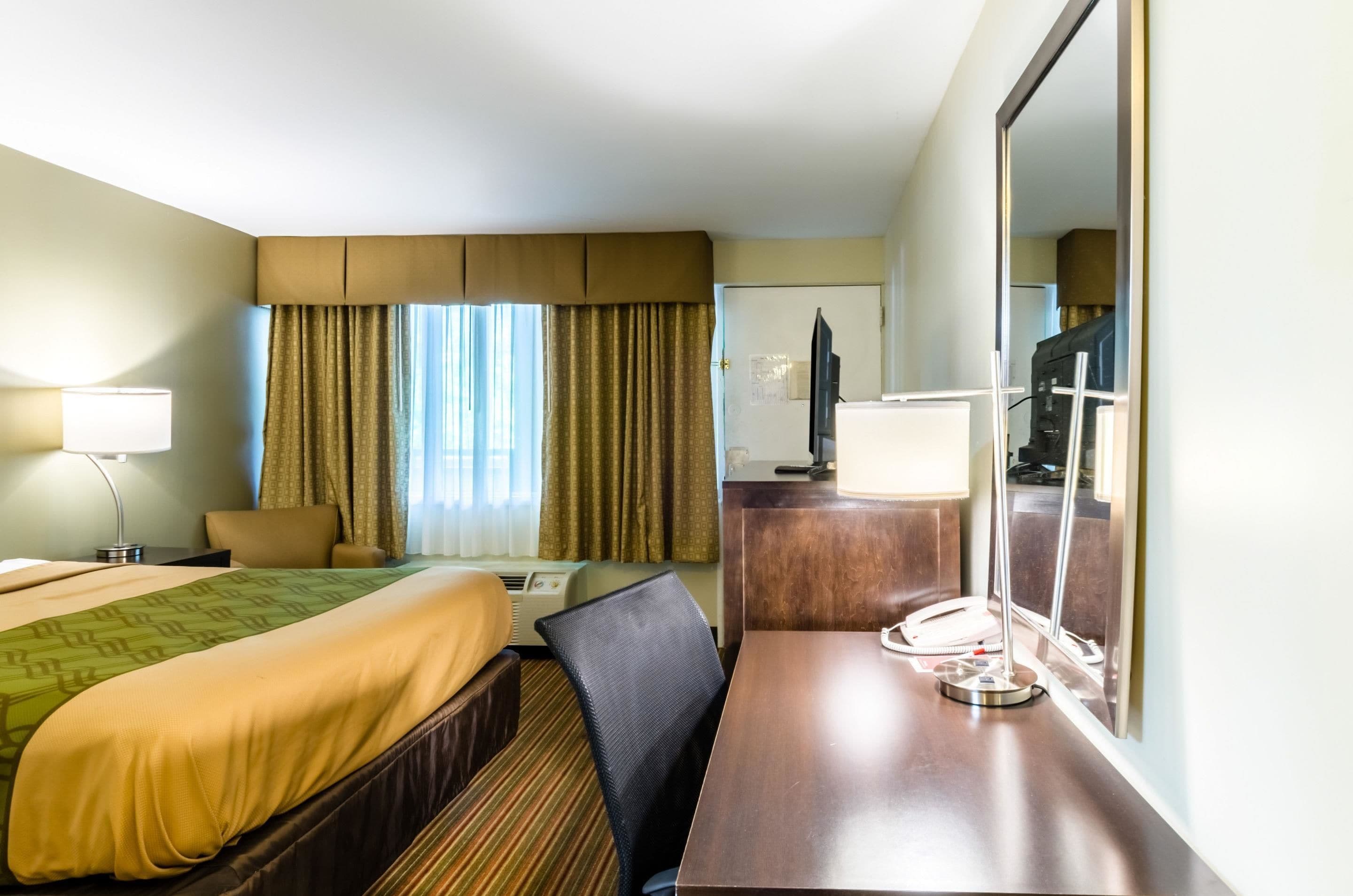 A hotel room with a bed, a desk, a chair, a desk lamp, a television, and a large mirror.