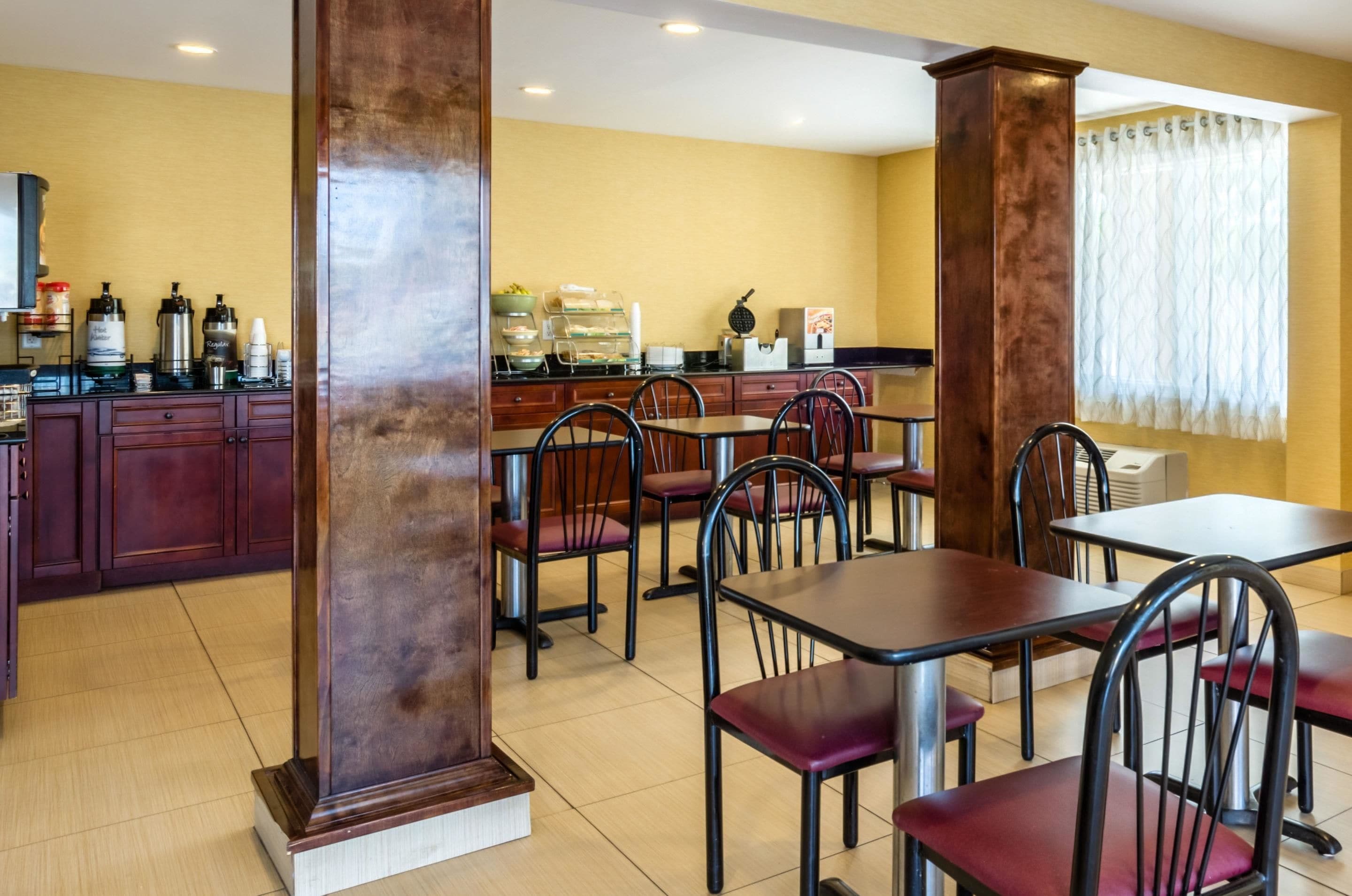 A hotel dining area with wooden tables and chairs, a coffee station with various coffee makers, and a yellow wall with a window.