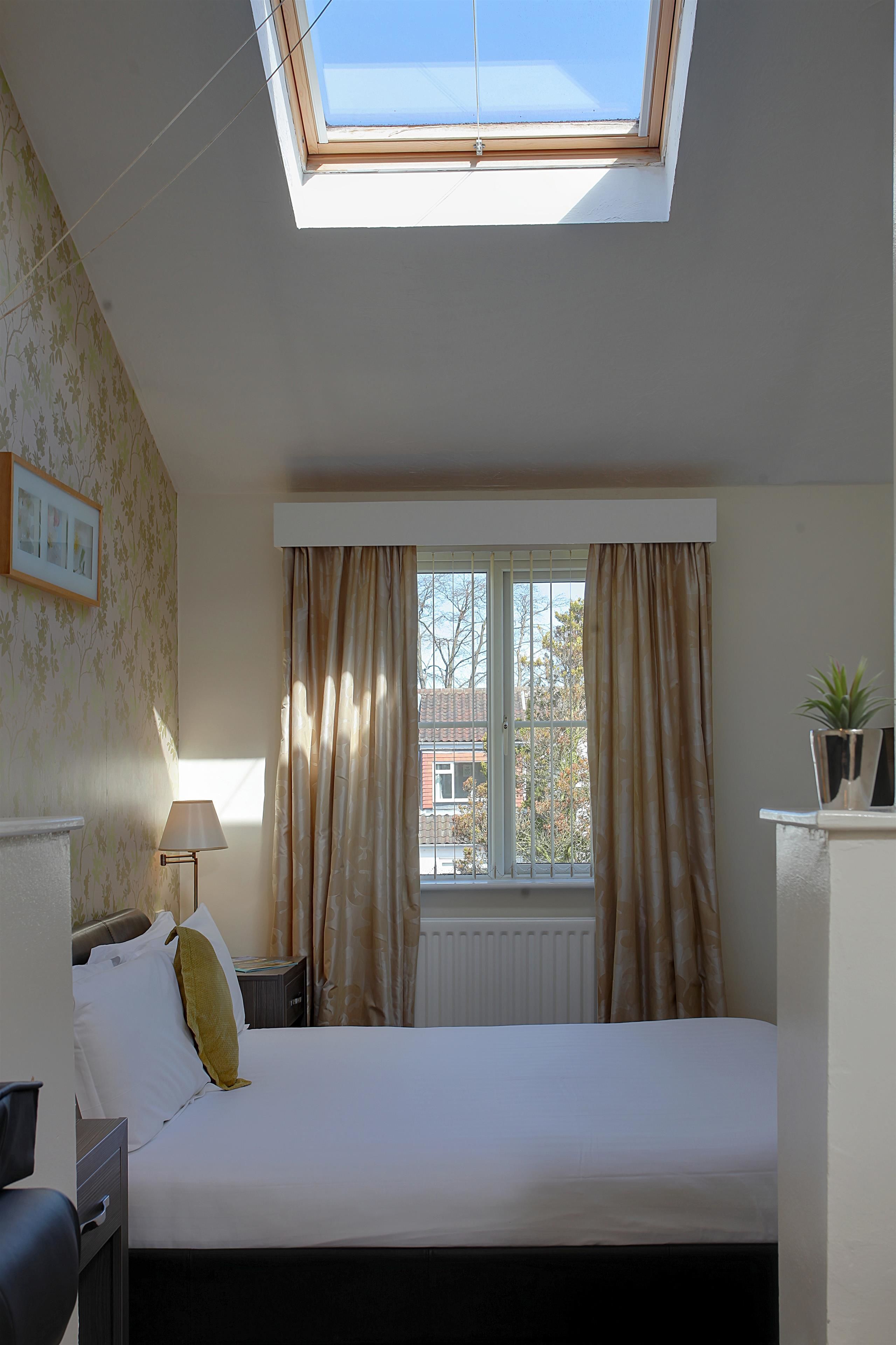 Ein Schlafzimmer mit Oberlicht, einem Bett, einem Nachttisch, einer Lampe und einem Fenster mit Vorhängen.