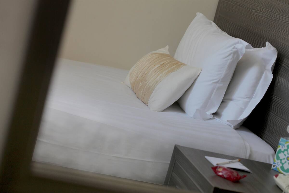 Un lit bien fait, avec du linge de lit blanc et un oreiller beige, une table de chevet avec une boîte de mouchoirs, un objet rouge et un objet blanc.