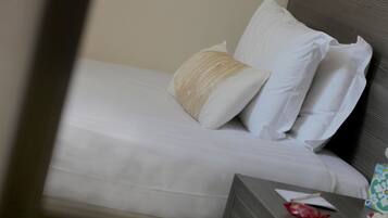 Un letto rifatto con lenzuola bianche e un cuscino beige, un comodino con una scatola di fazzoletti, un oggetto rosso e un oggetto bianco.