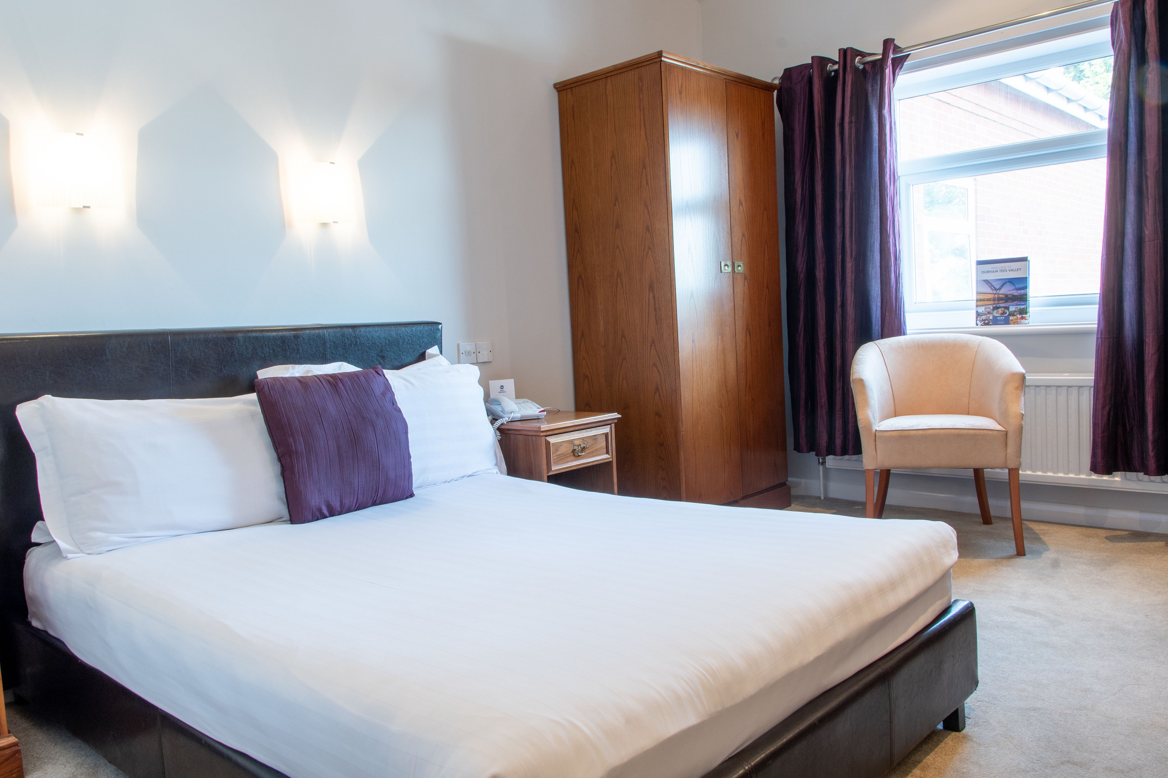 A hotel room with a large bed, a wardrobe, a chair, and a window with curtains.
