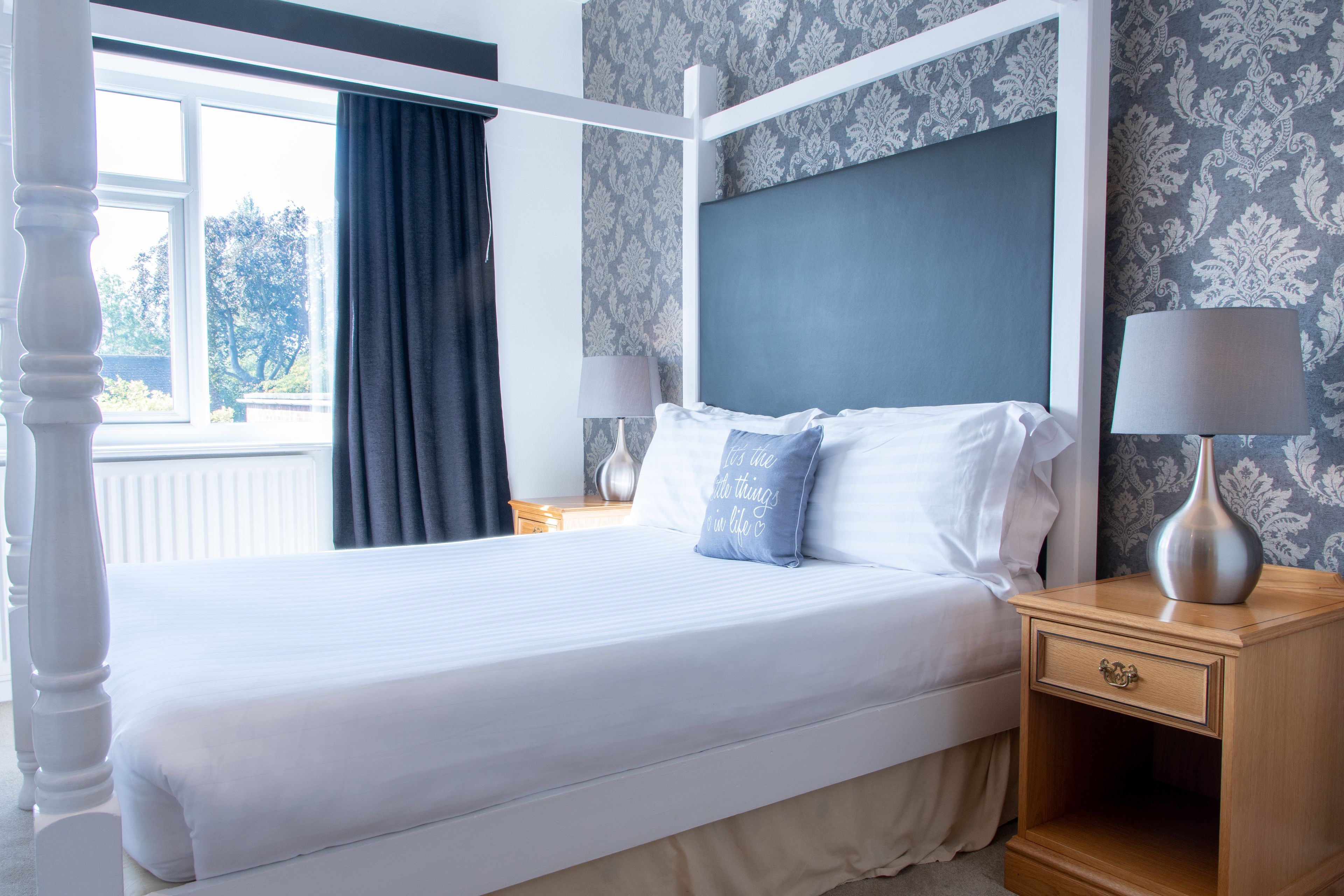 Una cama bien hecha con sábanas blancas y una almohada azul, una mesita de noche con una lámpara, y una ventana con cortinas.