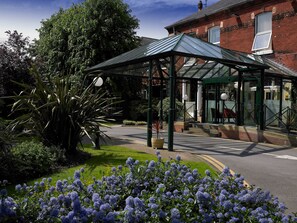 A covered outdoor area with a glass roof, surrounded by greenery and a pathway leading to a building entrance.