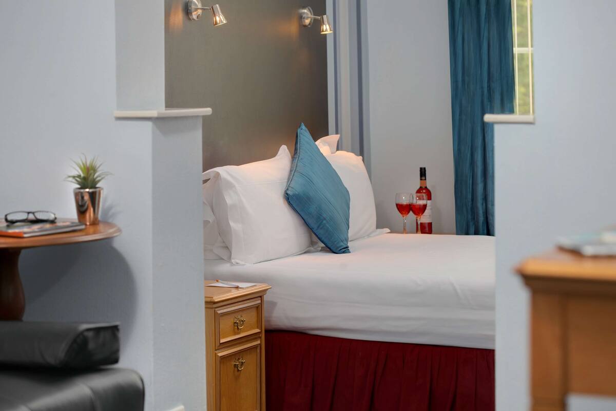 Une chambre d’hôtel avec un lit, une table de chevet, une petite table avec une plante, une bouteille de vin et une fenêtre avec des rideaux bleus.