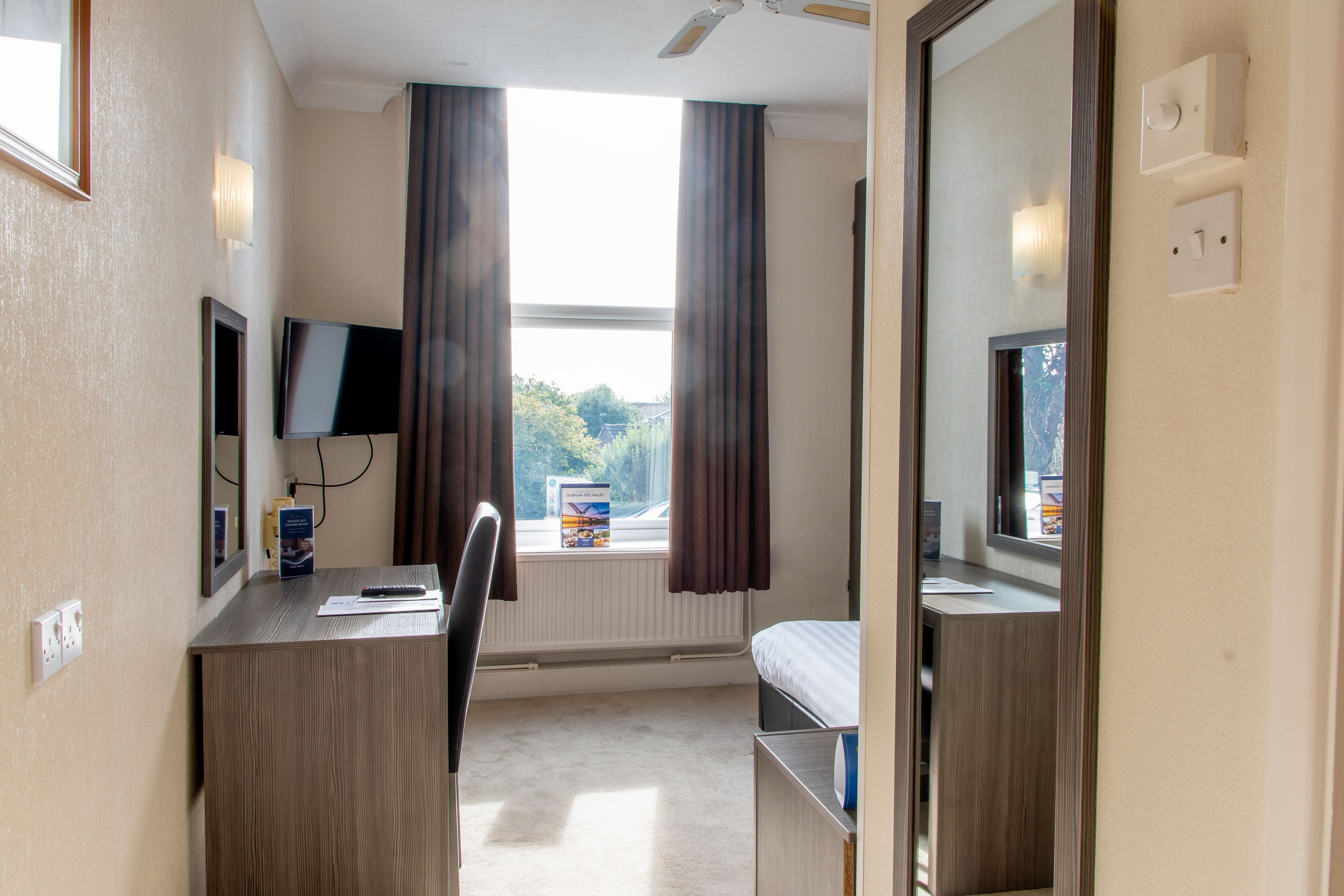 A hotel room with a desk, chair, and a bed. There is a window with curtains, a flat-screen TV mounted on the wall, and a mirror on the door.