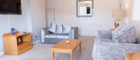 A living room with a flat-screen TV, a wooden coffee table, two grey sofas, a wooden side table, and a lamp.