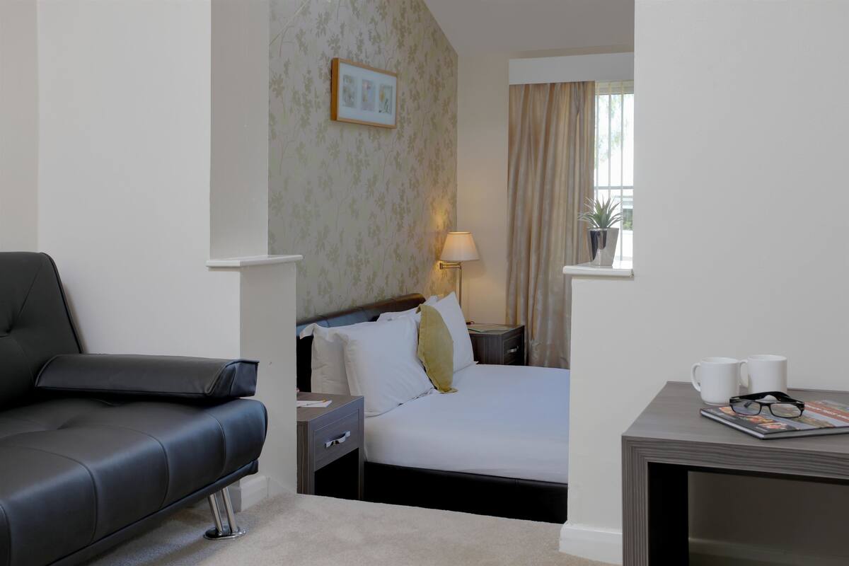 Une chambre d’hôtel avec un lit, une table de chevet, une lampe, une fenêtre avec des rideaux, une petite table avec un magazine et des verres, et un canapé en cuir noir.