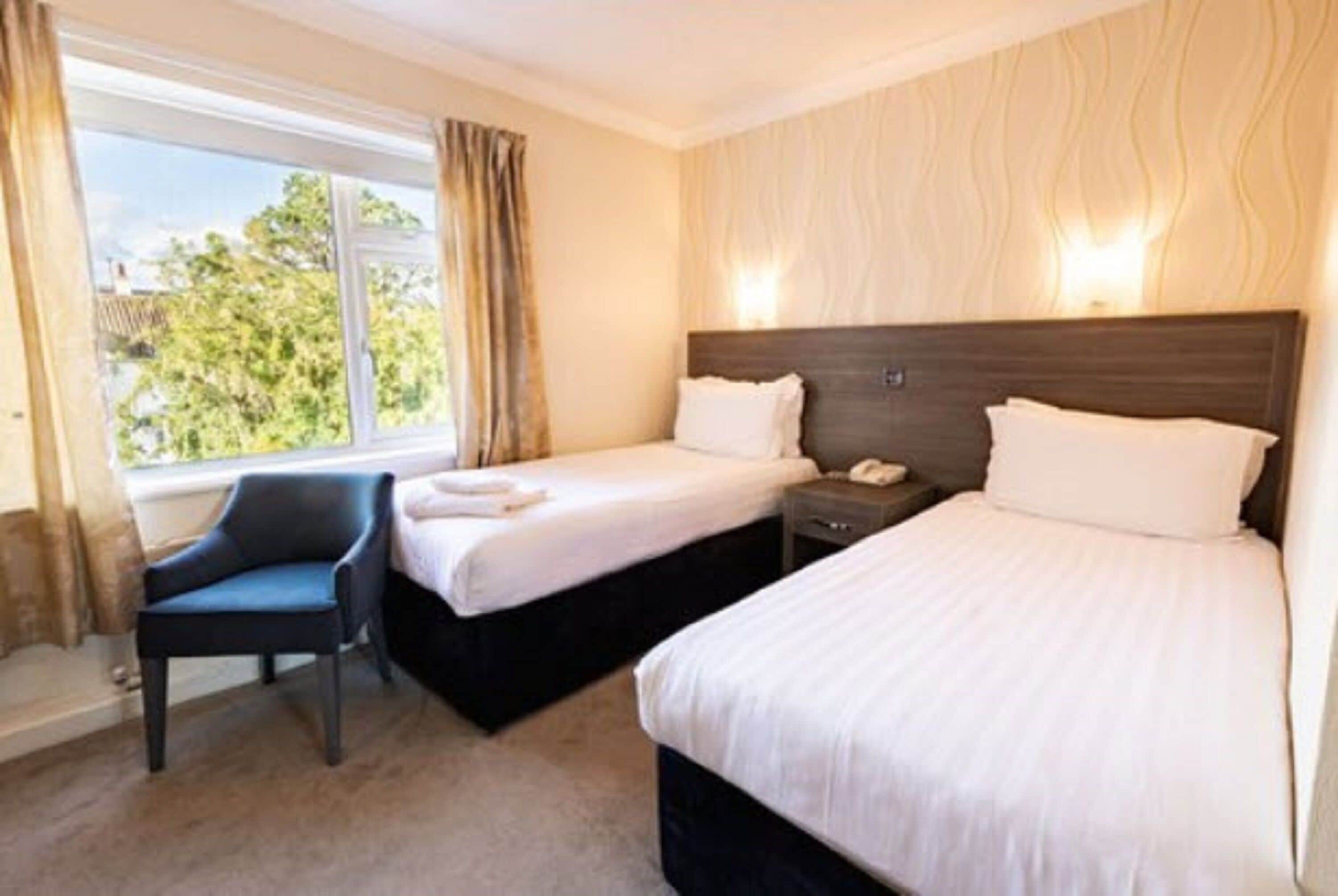 Quarto de hotel com duas camas de solteiro, uma cadeira, mesa de cabeceira com telefone e janela com cortinas.