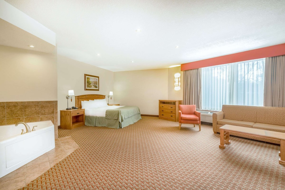 A hotel room with a large bed, a sitting area with a sofa and armchair, a small desk, a bathroom with a bathtub, and a large window with curtains.