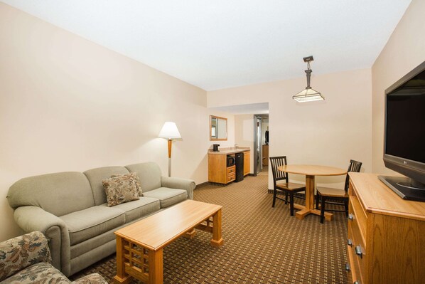 A compact living room with a sofa, a small table, a TV, and a kitchenette in the background. - Ramada Tropics Resort & Conf Center by Wyndham Des Moines (Urbandale)