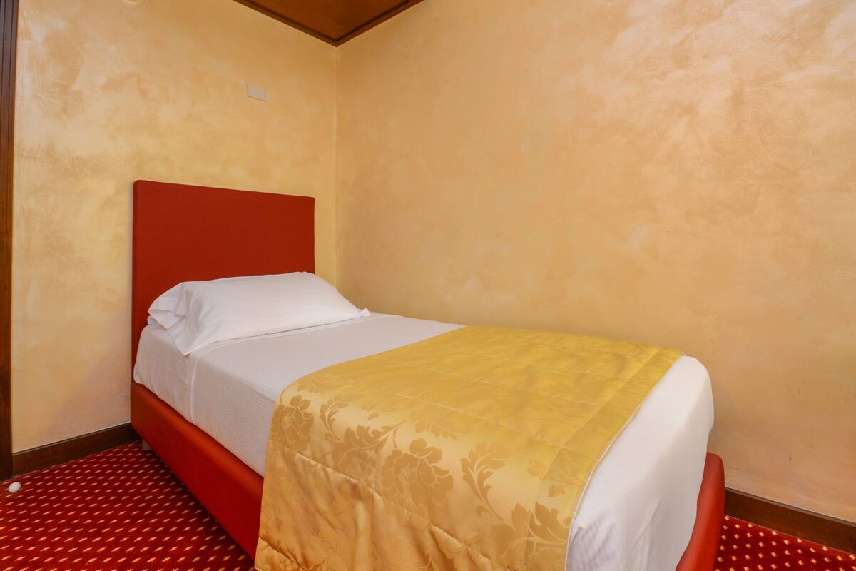 A single bed with a red headboard and a yellow bedspread in a room with beige walls and a red carpet.