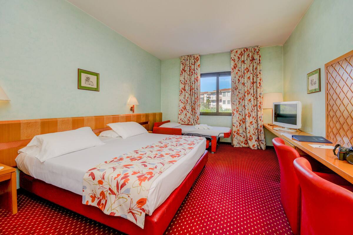 A hotel room with a large bed, a desk with a computer, a TV, and a window with curtains.