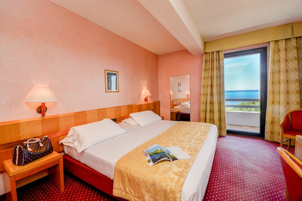 A hotel room with a bed, a nightstand, a chair, a desk, and a window with a view.