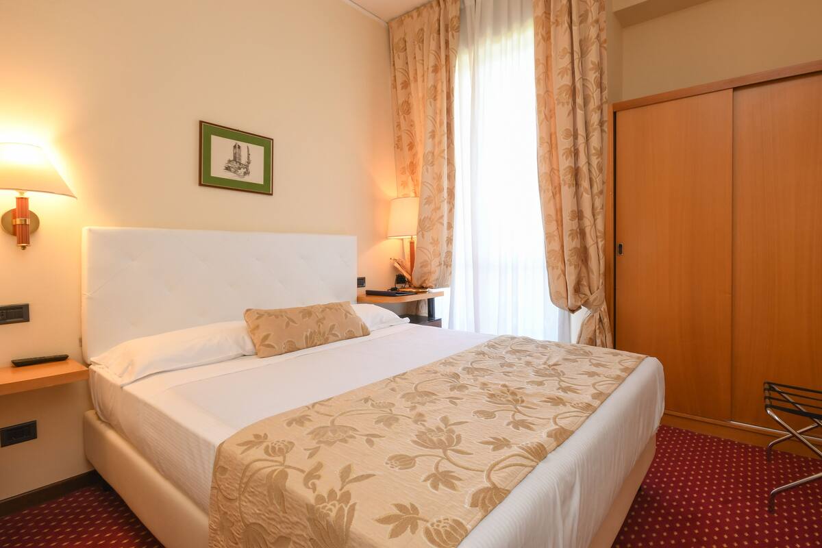 A hotel room with a large bed, a nightstand, a lamp, a framed picture on the wall, and a wardrobe.
