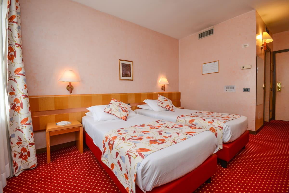 A hotel room with two beds, a wooden headboard, a nightstand, a red carpet, and two wall-mounted lamps.